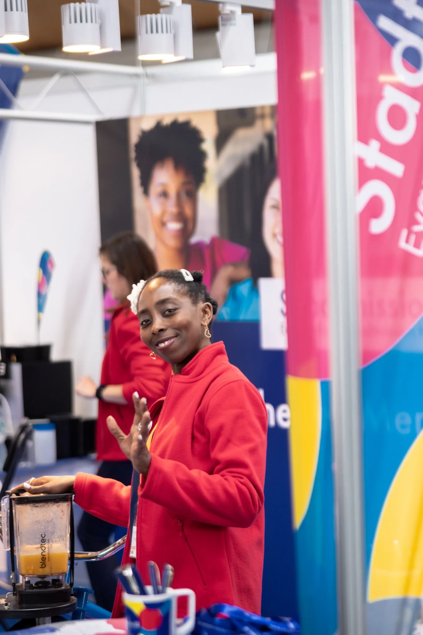 Lächelnde Frau im roten Mantel, die an einem Stand der Evangelischen Stadtmission mit einem Mixer arbeitet und der Kamera zuwinkt. Die Szene zeigt die freundliche Atmosphäre bei einer Veranstaltung der Sozialstation Karlsruhe, mit den Themen Fotografie, Social Media, Videografie und Konzept.