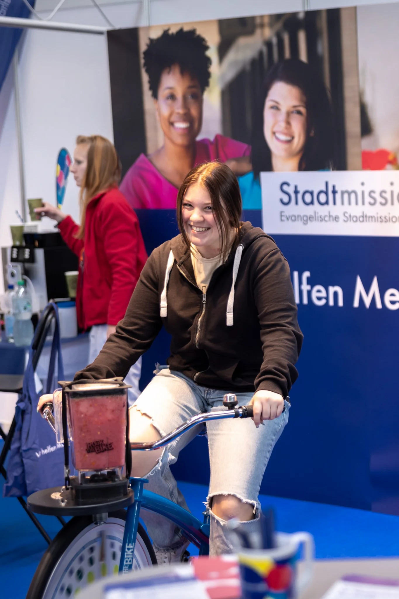 Junge Frau auf einem Standfahrrad mit integriertem Mixer bei einer Veranstaltung der Evangelischen Stadtmission. Sie lächelt während sie das Rad fährt, das für das Mixen eines Getränks genutzt wird. Dieses Bild repräsentiert die Sozialstation Karlsruhe mit den Themen Fotografie, Social Media, Videografie und Konzept.