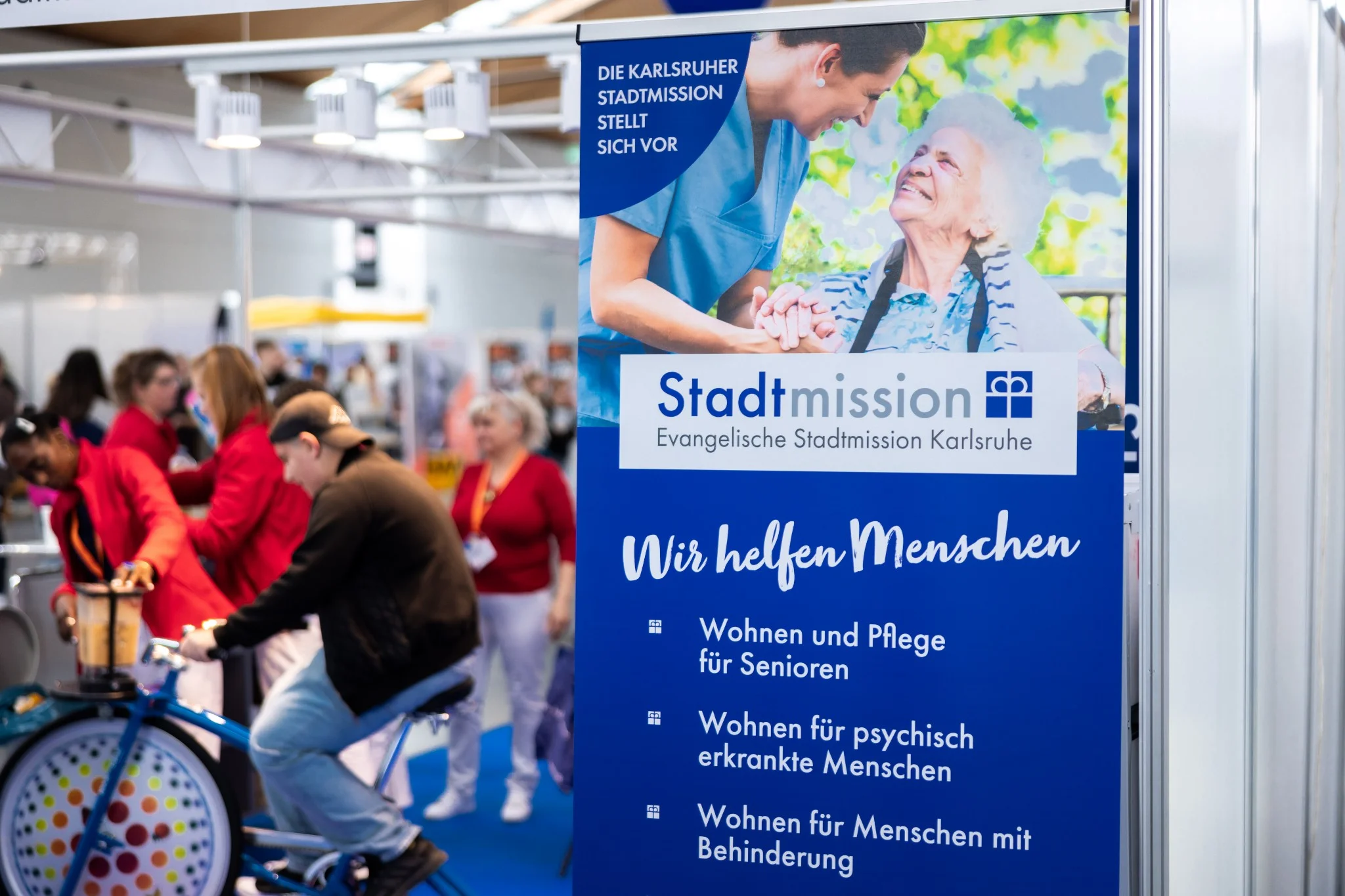 Informationsstand der Stadtmission Karlsruhe auf einer Messe, mit einem Banner, das über die Angebote der Evangelischen Stadtmission Karlsruhe informiert: Wohn- und Pflegeangebote für Senioren, Wohnangebote für psychisch erkrankte Menschen und Menschen mit Behinderung.