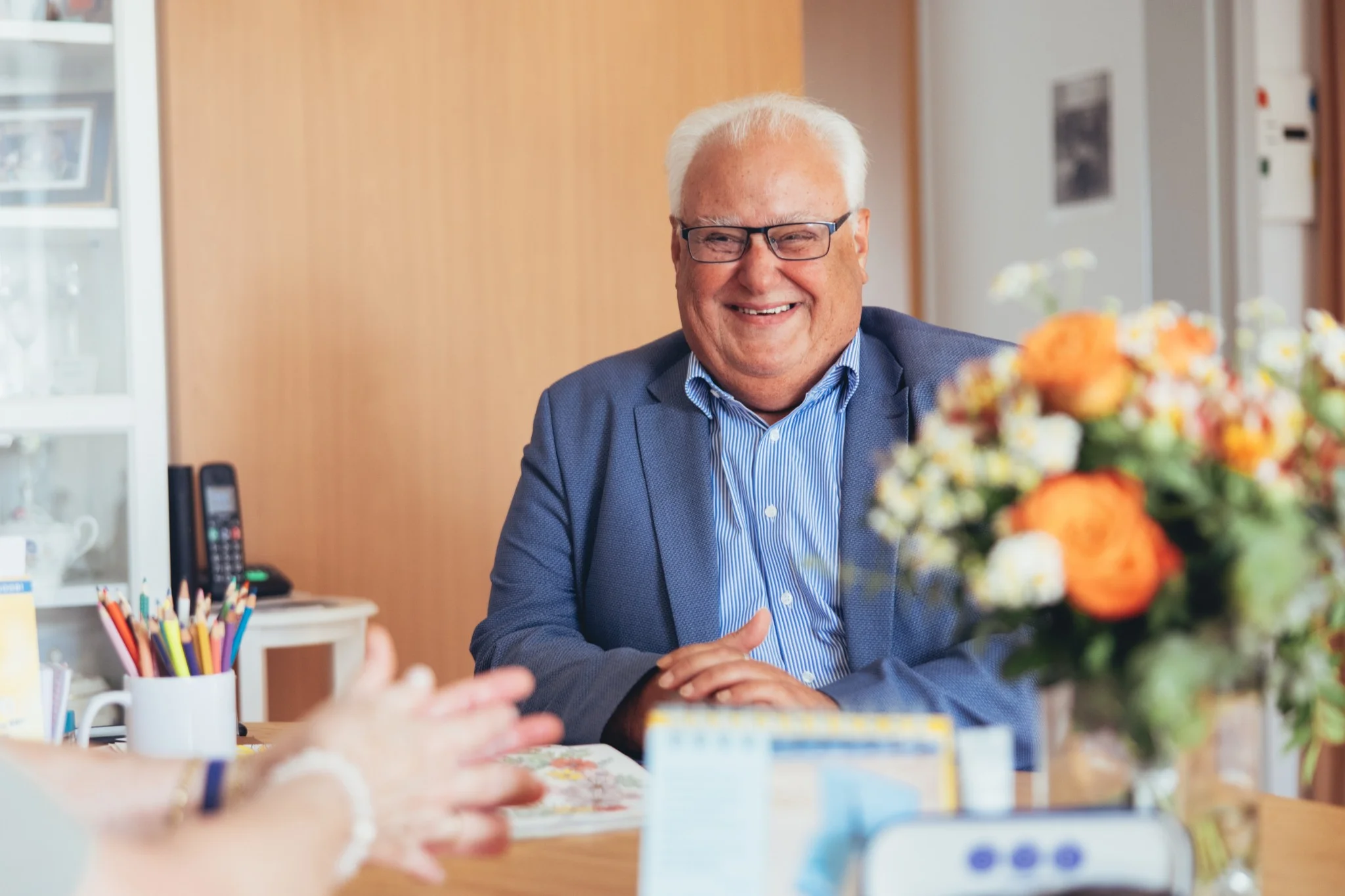 Freundlicher älterer Herr im Gespräch in einem Büro der Sozialstation Karlsruhe, umgeben von Blumen und Schreibmaterial. Syd.media unterstützt die Sozialstation mit Projektbegleitung und Fotografie für ihre Kommunikationsarbeit.