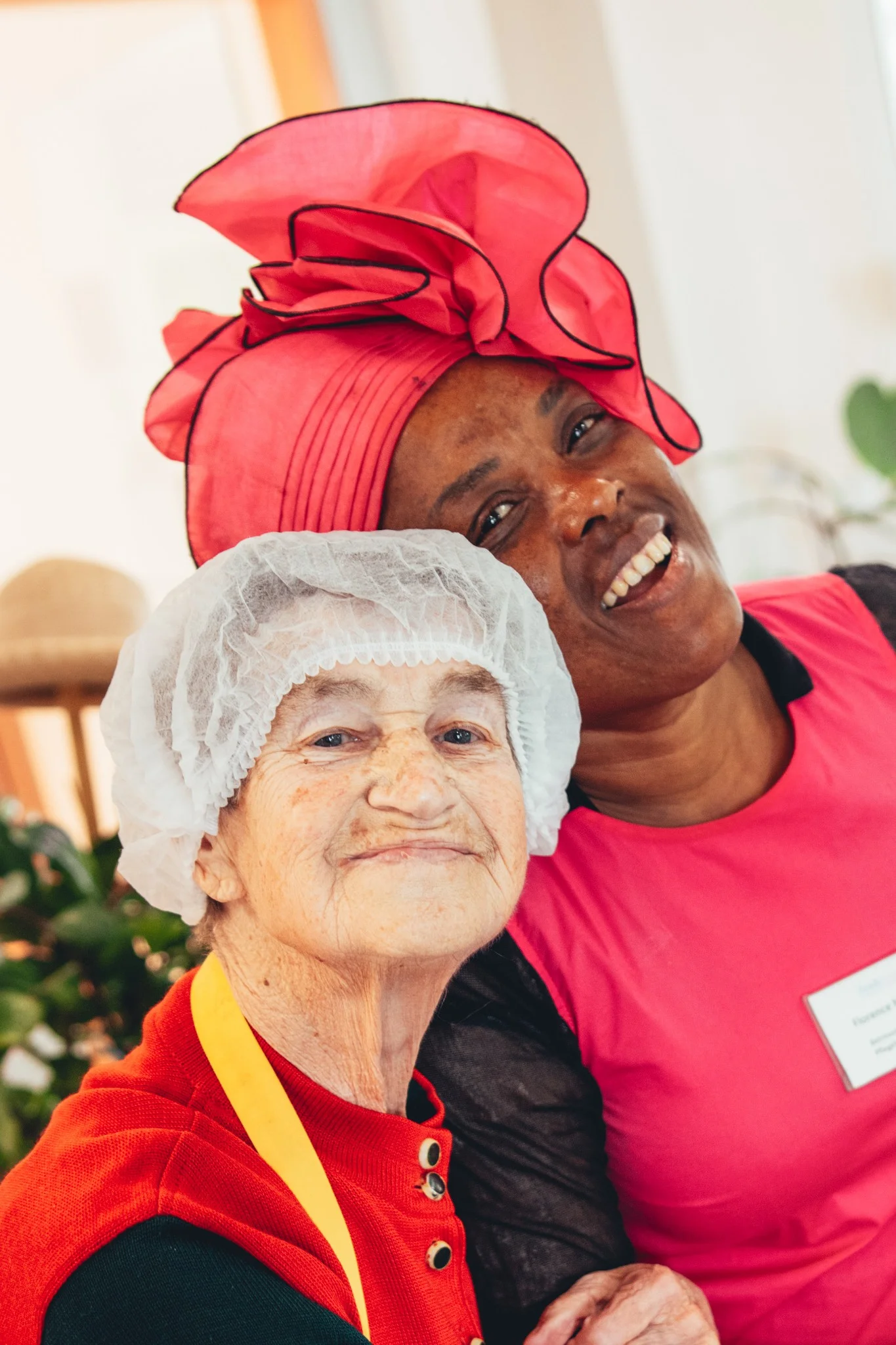 Fröhliche Seniorin mit Haarnetz und Pflegekraft mit leuchtend pinkem Kopftuch bei einer Gemeinschaftsaktivität in der Sozialstation Karlsruhe, unterstützt durch das Team von syd.media und die Stadtmission.