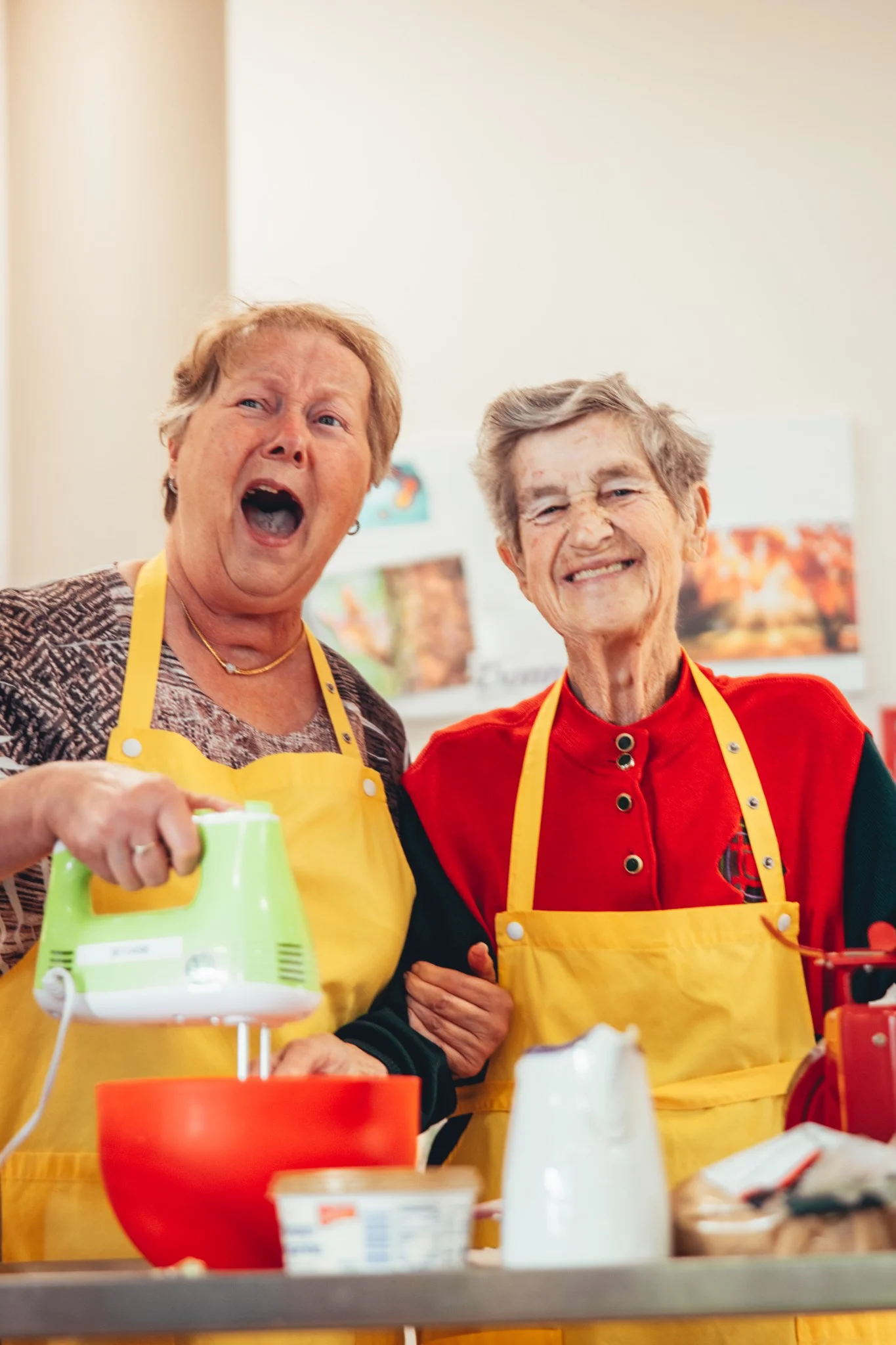 Zwei fröhliche Seniorinnen in gelben Schürzen bei einer gemeinsamen Backaktivität in der Sozialstation Karlsruhe, unterstützt durch das Team von syd.media für die Stadtmission.