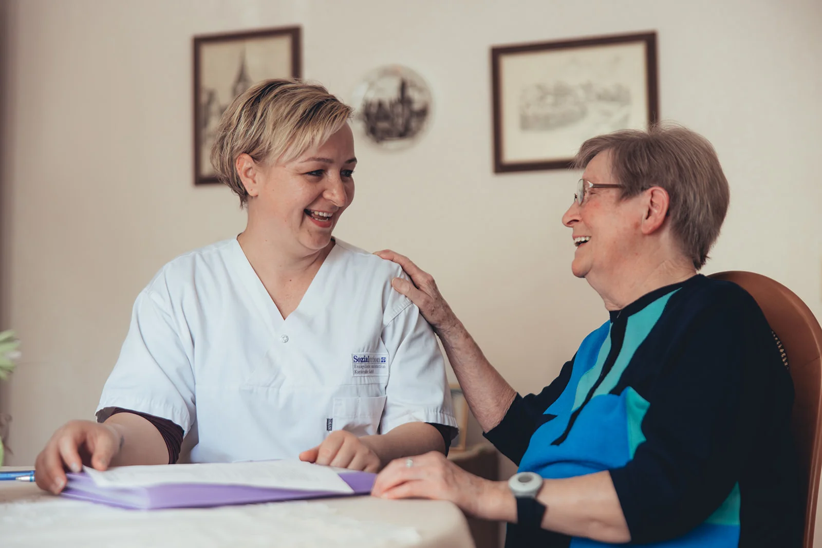 Eine Pflegekraft der Sozialstation in freundlichem Gespräch mit einer älteren Dame am Tisch, beide lachen miteinander. Die Szene vermittelt Nähe und Vertrauen und repräsentiert die wertvolle Arbeit der Sozialstation in Karlsruhe.