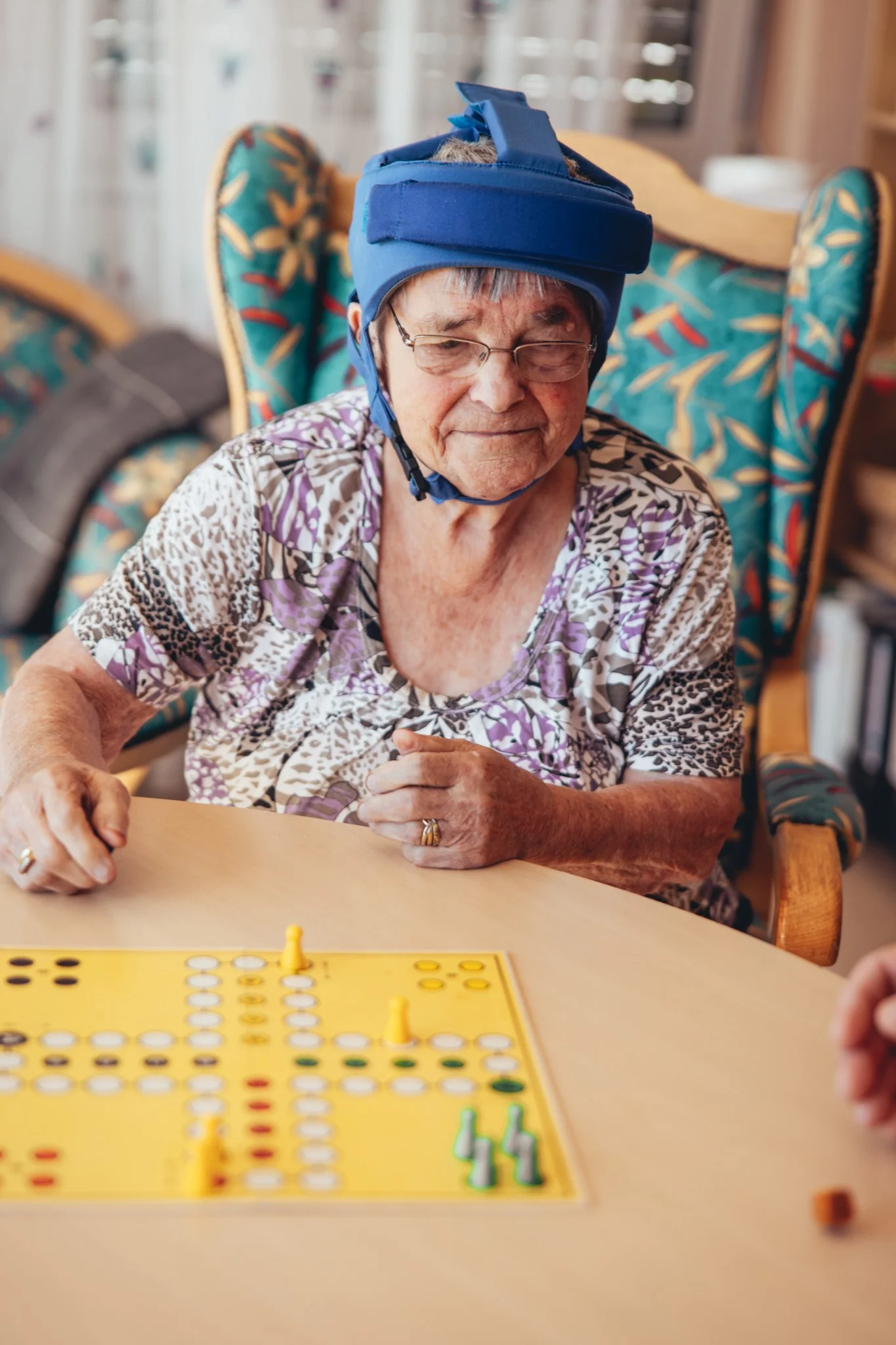 Seniorin mit Schutzhelm beim Gesellschaftsspiel im Betreuungsbereich der Stadtmission Karlsruhe. Spielerische Aktivität zur Förderung der sozialen Interaktion und Betreuung von Senioren.